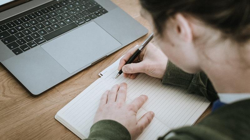 An image of a woman writing in a notebook with a laptop in the background